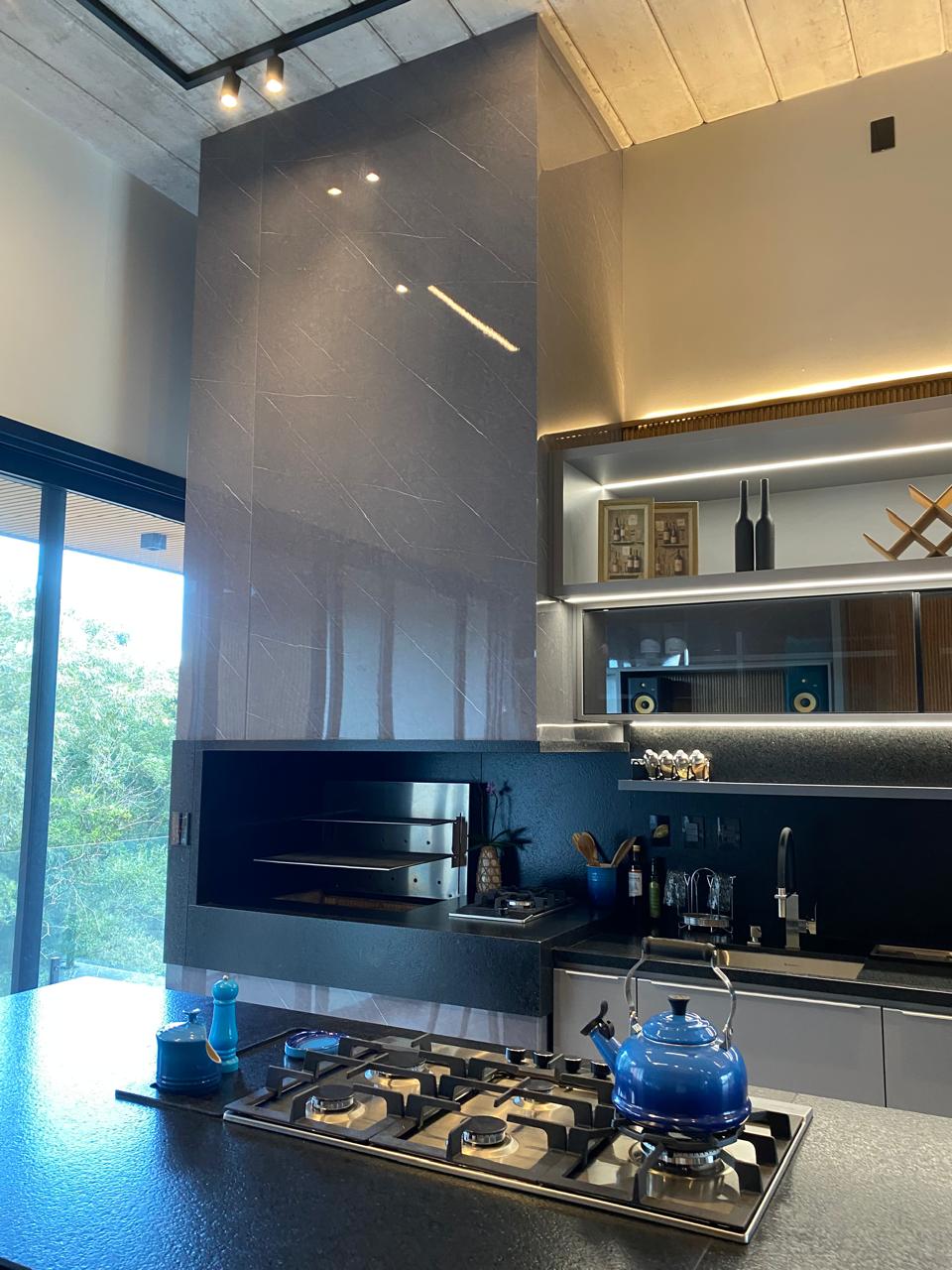 Área de churrasqueira gourmet com parede revestida em porcelanato grafite marmorizado, churrasqueira embutida em concreto escuro, bancada em granito preto, armários planejados em madeira clara, fogão cooktop e ambiente integrado com jardim, fotografia de interiores realista