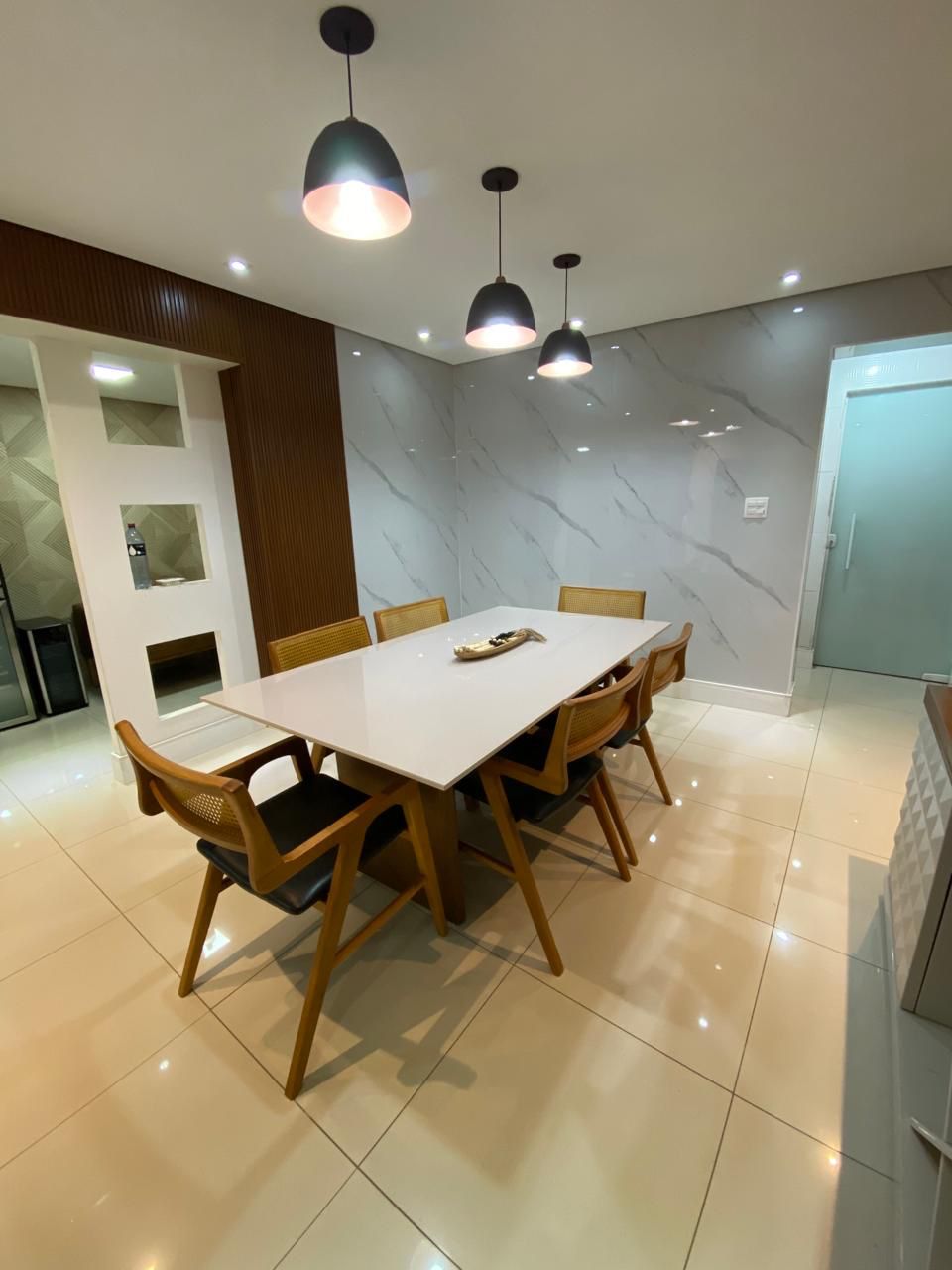 Sala de jantar moderna com piso porcelanato bege acetinado de alto brilho, parede em mármore branco e dourado, mesa retangular branca com cadeiras de madeira e palhinha, luminárias pendentes pretas, iluminação aconchegante, fotografia de interiores realista