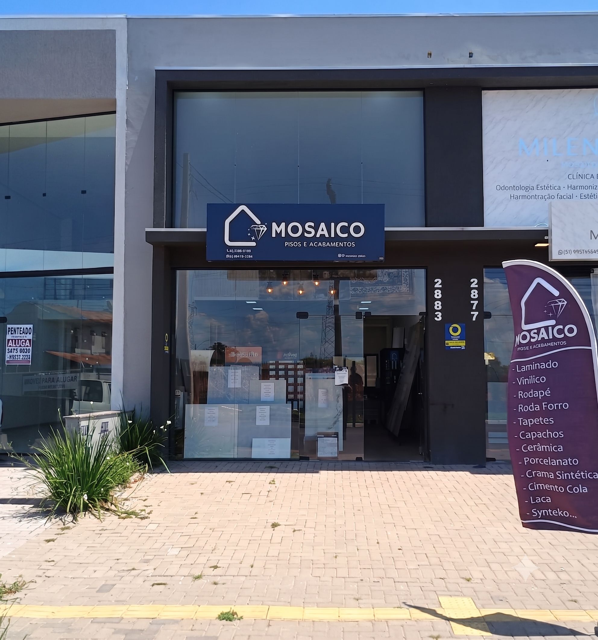 Fachada da loja Mosaico Pisos e Acabamentos em Canoas, com letreiro azul-marinho e logotipo, vitrine de vidro exibindo amostras de pisos e revestimentos, banner roxo com lista de produtos, dia ensolarado com céu azul, fotografia comercial realista